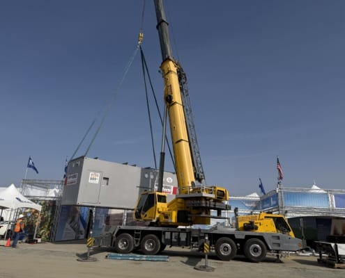 A large yellow Grove mobile crane lifting a grey BSD Builders modular microgrid unit into place at the World Ag Expo under a clear blue sky.