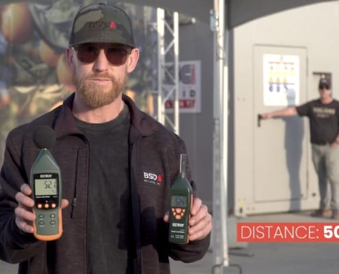 A BSD Builders team member holding two Extech sound level meters showing decibel readings of 52.1 and 52.4 at a distance of 50 feet from the SSC Microgrid unit during a field test in Tulare, CA.